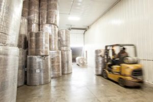 Unfaced wide-roll insulation blankets rolled and wrapped in plastic, stacked high on one side of a warehouse room.