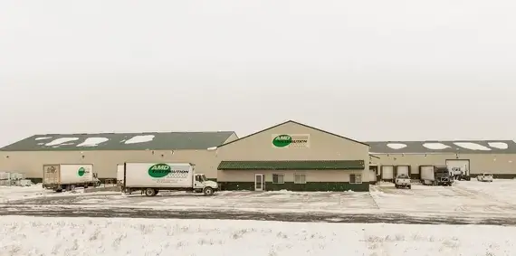 Long, one-story beige metal AMD Distribution building with large mostly-empty parking lot in front, with light dusting of snow and white/gray sky behind.