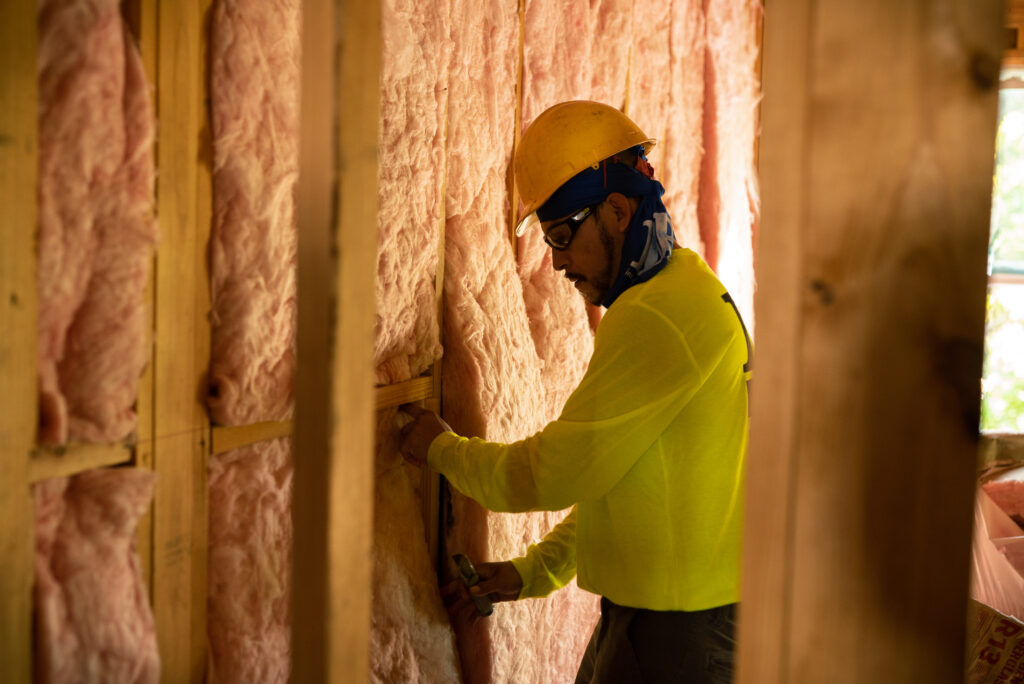 Technician in yellow shirt and hardhat, installing batt fiberglass insulation in a wall.